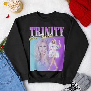 Könnte beinhalten: Schwarzer Pullover mit einem Grafikdruck von Trinity the Tuck aus RuPaul's Drag Race. Die Grafik ist ein Foto im Vintage-Stil von Trinity vor einem rosa und blauen Hintergrund mit dem Text "TRINITY the Tuck" in einem lila und rosa Farbverlauf.