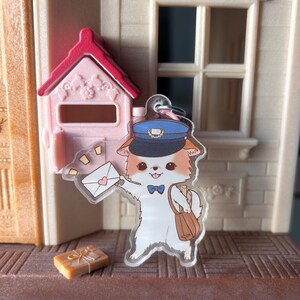 May include: A clear acrylic charm depicting a cartoon cat postman, holding an envelope with a heart. The cat wears a blue hat and carries a brown satchel. A pink mailbox and a small gift box are also visible.