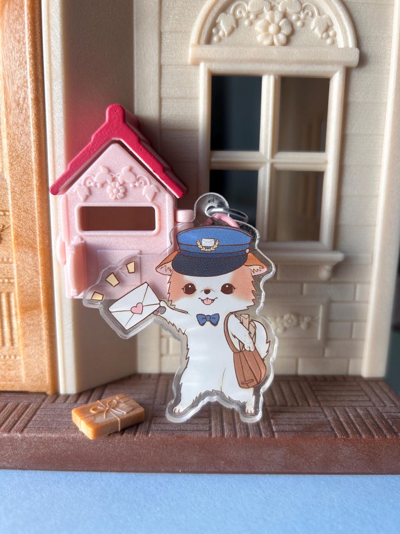 May include: A clear acrylic charm depicting a cartoon cat postman, holding an envelope with a heart. The cat wears a blue hat and carries a brown satchel. A pink mailbox and a small gift box are also visible.