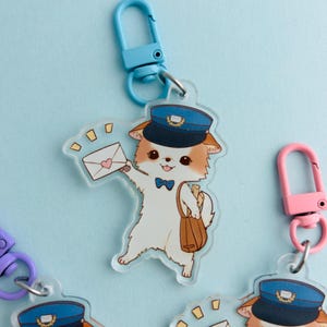 May include: A clear acrylic keyring featuring a cartoon dog dressed as a postman, wearing a blue hat and bow tie, holding an envelope. The keyring has a blue clasp and a brown satchel. Other keyrings with different coloured clasps are also visible.