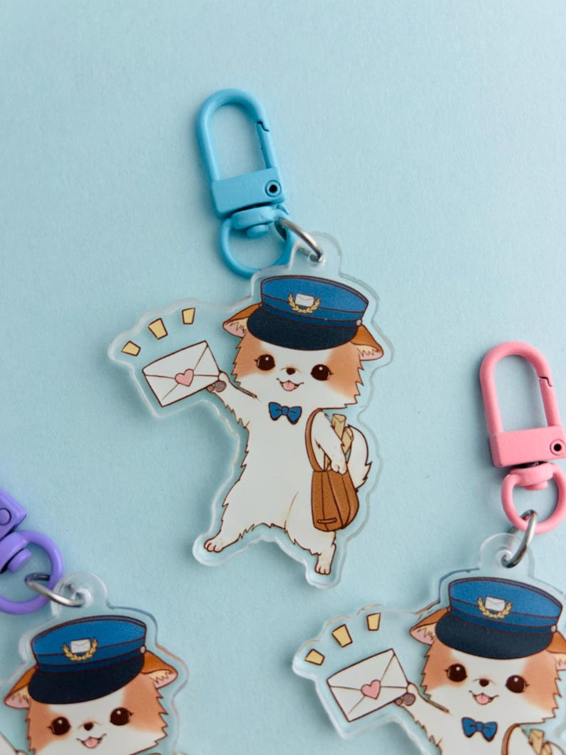 May include: A clear acrylic keyring featuring a cartoon dog dressed as a postman, wearing a blue hat and bow tie, holding an envelope. The keyring has a blue clasp and a brown satchel. Other keyrings with different coloured clasps are also visible.