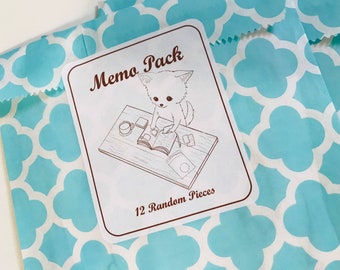 May include: A pale blue paper bag with a white floral design. A white label on the bag reads "Memo Pack" and features a cartoon dog at a desk. The label also says "12 Random Pieces".