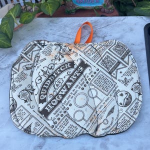 May include: A quilted fabric pot holder featuring a cream-colored background with black text and illustrations related to Quidditch at Hogwarts. The design includes diagrams of hoops, a Golden Snitch, and a Bludger, with an orange fabric hanging loop.
