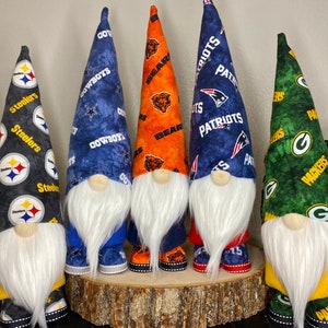 May include: Five decorative gnomes with long, pointed hats and white beards. Each gnome's hat features a different team's logo and colors, including Steelers, Cowboys, Bears, Patriots, and Packers. The gnomes are displayed on a wooden base.