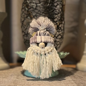 May include: A white and gray gnome with a white and gray striped hat and a white beard. The gnome is wearing a small white flower on its hat.