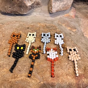 May include: A collection of eight cat-shaped ornaments crafted from small plastic pony beads. Each figure features a distinct color pattern, including solid black, brown, white, light blue, tan, and multi-colored striped designs with long tails.
