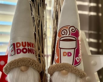 Könnte beinhalten: Zwei Zwergfiguren mit weißen Kegelhüten mit dem Dunkin' Donuts-Logo. Die Zwerge haben lange weiße Bärte, einen beigefarbenen Pelzbesatz und rosa und braune Kleidung. Der Hintergrund umfasst getrocknete Pflanzen und eine sternförmige Dekoration.