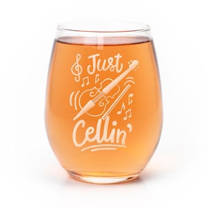 May include: A stemless wine glass filled with amber liquid, featuring a white etched graphic of a cello, a bow, musical notes, and the text "& Just Cellin'". The design is centered on the front of the clear glass, showcasing a musical theme.