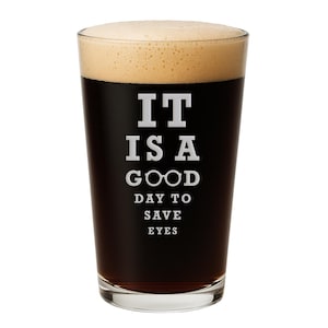 May include: A clear glass pint beer glass filled with a dark brown liquid topped with a thick tan foam head. The glass features white printed text that reads IT IS A GOOD DAY TO SAVE EYES, with the two O's in GOOD replaced by a pair of eyeglasses.