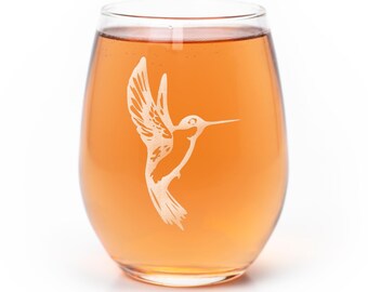 Könnte beinhalten: Ein klares, stieloses Glas, gefüllt mit einer bernsteinfarbenen Flüssigkeit. Auf der Vorderseite des Glases befindet sich eine weiße, geätzte Grafik eines fliegenden Kolibris mit nach oben ausgebreiteten Flügeln und einem langen, dünnen Schnabel.