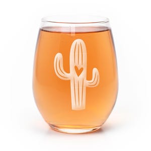 May include: A clear stemless wine glass filled with a light orange liquid. The glass features a white etched graphic of a saguaro cactus with a small heart shape cut out from the center of the cactus body, centered on the front of the glass.