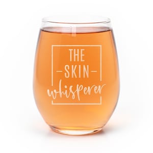 May include: A stemless glass filled with an amber-colored liquid. The glass features a white etched rectangular frame containing the text THE -SKIN- whisperer in a combination of clean sans-serif and elegant cursive script lettering.