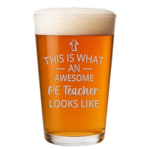May include: A clear glass pint beer mug filled with amber-colored beer and topped with white foam. The glass features an upward-pointing arrow and the white etched text: THIS IS WHAT AN AWESOME PE Teacher LOOKS LIKE, centered on the front surface.