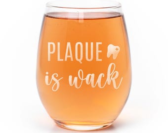 May include: A stemless wine glass filled with an amber-colored liquid. The glass features white etched text that reads PLAQUE is wack, accompanied by a small graphic of a tooth with a four-pointed star sparkle on its surface.