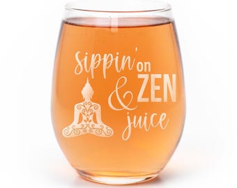 Può includere: Un calice da vino senza stelo trasparente riempito con un liquido color ambra chiaro. Il bicchiere presenta una scritta incisa in bianco che recita sippin' on & ZEN juice, accompagnata da un grafico bianco dettagliato di una figura meditante.