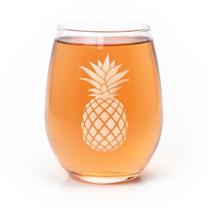 May include: A clear, stemless glass filled with a light orange liquid. The front of the glass features a white, etched pineapple design centered on the surface. The glass has a rounded, bulbous shape with a smooth, curved rim and a solid base.