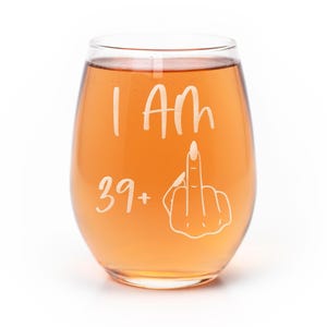 May include: A clear stemless wine glass filled with an amber-colored liquid. The glass features white etched text that reads I Am 39+ next to a graphic illustration of a hand making a middle finger gesture with long manicured fingernails.