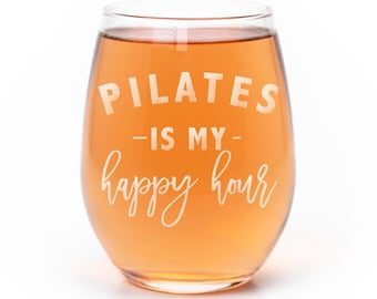 May include: A stemless wine glass filled with a light orange liquid. The glass features white etched text that reads PILATES -IS MY- happy hour, with the word PILATES in a bold sans-serif font and happy hour in a cursive script font.