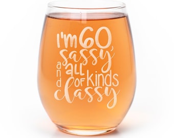 May include: A clear stemless wine glass filled with a light orange liquid. The glass features white etched text that reads: I'm 60, sassy, and all kinds of classy. The design is centered on the front of the glass with a mix of block and script fonts.
