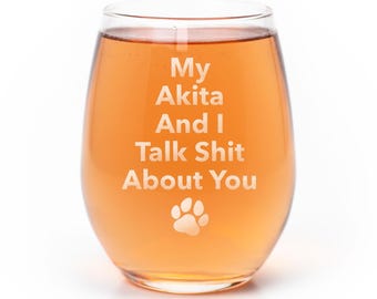 May include: A clear stemless wine glass filled with an amber-colored liquid. The glass features white etched text that reads My Akita And I Talk Shit About You, positioned above a small graphic of a dog paw print centered near the bottom.
