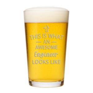 May include: A clear glass pint beer mug filled with golden lager and a thick white foam head. The glass features an upward-pointing arrow and the etched text: THIS IS WHAT AN AWESOME Engineer LOOKS LIKE, centered on the front surface of the glass.