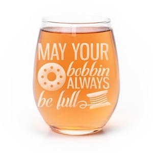 May include: A stemless wine glass filled with an amber-colored liquid. The glass features white etched text that reads MAY YOUR bobbin ALWAYS be full, accompanied by a graphic of a sewing bobbin and a spool of thread with a trailing line.