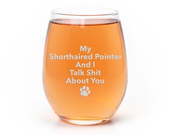 May include: A stemless wine glass filled with an orange liquid. The glass has the text "My Shorthaired Pointer And I Talk Shit About You" and a paw print graphic etched on the front. The glass is clear and sits on a white background.
