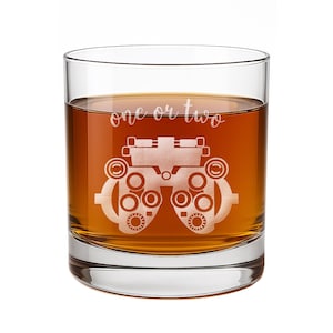 May include: A clear glass tumbler filled with an amber-colored liquid. The front of the glass features an etched graphic of an optometry phoropter and the white script text one or two, positioned directly above the medical eye testing device.