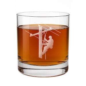 May include: A clear glass tumbler filled with amber liquid, featuring a white etched graphic of a utility worker climbing a wooden power pole with electrical lines extending horizontally across the center of the glass surface.