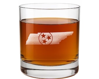 May include: A clear glass tumbler filled with amber-colored liquid, featuring an etched white silhouette of the state of Tennessee on the front. The state outline contains a circular emblem with three centered stars, consistent with the state flag.