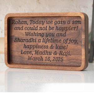 May include: A wooden tray with a personalized message engraved on it. The message reads: "Rohan, Today we gain a son and could not be happier! Wishing you and Sharadhi a lifetime of joy, happiness & love! Love, Madhu & Raji. March 16, 2025"