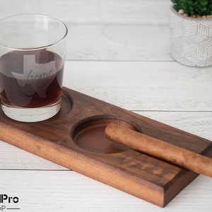 May include: A wooden tray with two circular indentations, one holding a glass of whiskey and the other a cigar. The glass has an etched design of the state of Texas with the word "home" inside. The tray is made of dark wood and has a natural finish.
