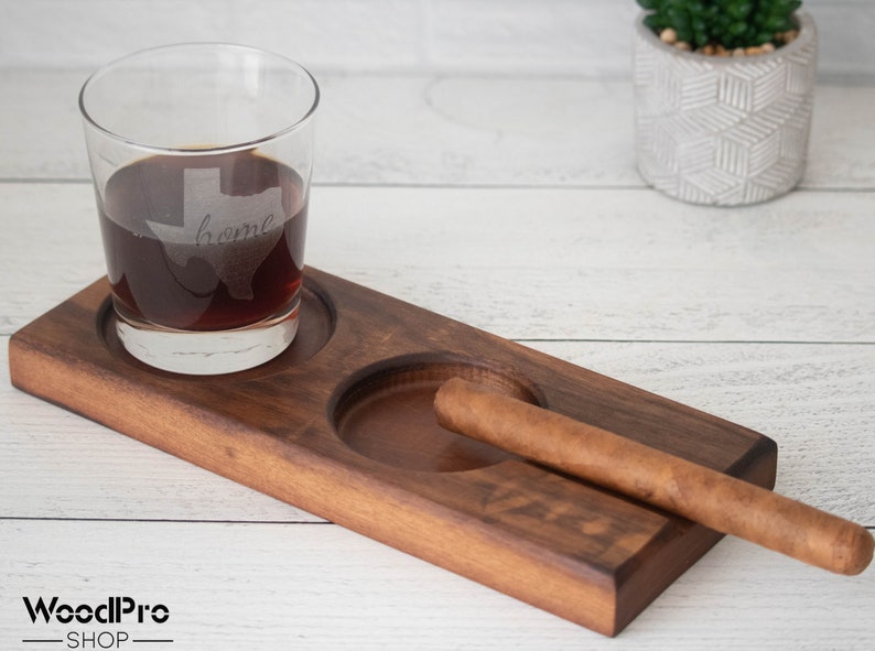 May include: A wooden tray with two circular indentations, one holding a glass of whiskey and the other a cigar. The glass has an etched design of the state of Texas with the word "home" inside. The tray is made of dark wood and has a natural finish.