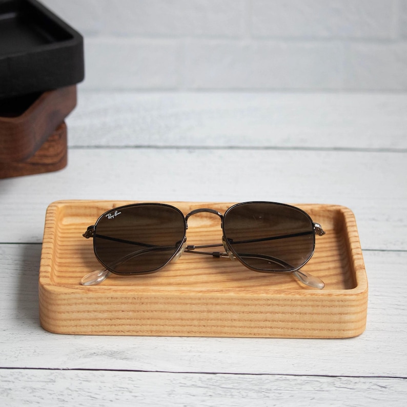 May include: A pair of dark-lensed sunglasses rests on a light-colored wooden tray. The tray is rectangular with raised edges. The sunglasses have a thin, dark metal frame. Other wooden trays are stacked in the background.