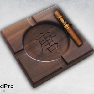 May include: A square wooden cigar ashtray with a round inset for ashes. The ashtray has a carved design of a Chinese character in the center. A brown cigar is resting on the ashtray.
