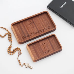 May include: Two wooden trays with a cross design and personalized engravings. The larger tray reads "Owen Manning Still" and the smaller tray reads "WJP". A wooden rosary with a silver cross is in the foreground.