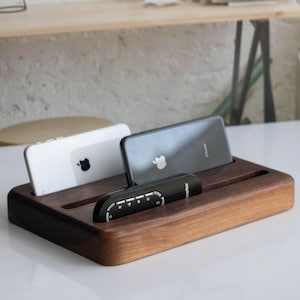 Puede incluir: Un soporte de madera para teléfono y mando a distancia con dos teléfonos inteligentes y un mando a distancia descansando en las ranuras. El soporte está hecho de madera de nogal oscuro.
