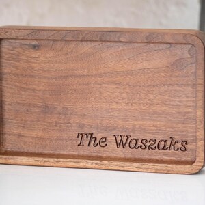 May include: A rectangular wooden tray with rounded corners. The tray is made of dark brown wood and has a recessed interior. The words "The Waszaks" are engraved on the tray's surface.