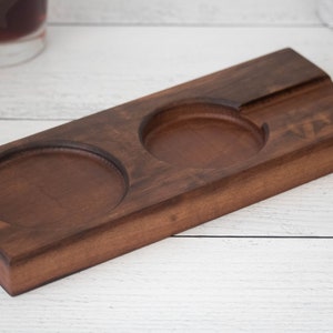 May include: A wooden coaster with two circular indentations for holding glasses and a rectangular indentation for holding a cigar. The coaster is made of dark brown wood and has the word "VIBES" engraved on it.