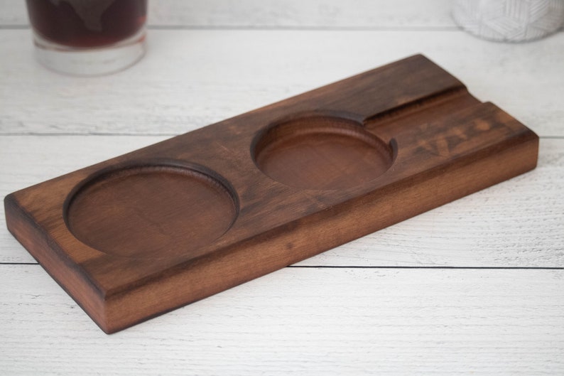 May include: A wooden coaster with two circular indentations for holding glasses and a rectangular indentation for holding a cigar. The coaster is made of dark brown wood and has the word "VIBES" engraved on it.