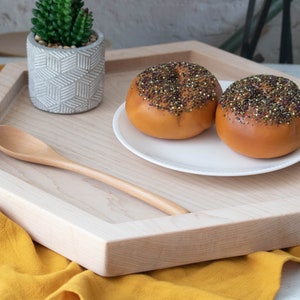 May include: A wooden hexagonal tray with a light wood finish. The tray has a small wooden spoon, a white ceramic planter with a succulent plant, and a white plate with two round buns topped with black and gold sprinkles.