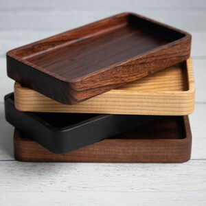 May include: A stack of four rectangular wooden trays in varying shades of brown and black. The top tray is a dark, rich brown, followed by a lighter wood, a black tray, and a medium brown tray at the bottom. The trays are stacked on top of each other.