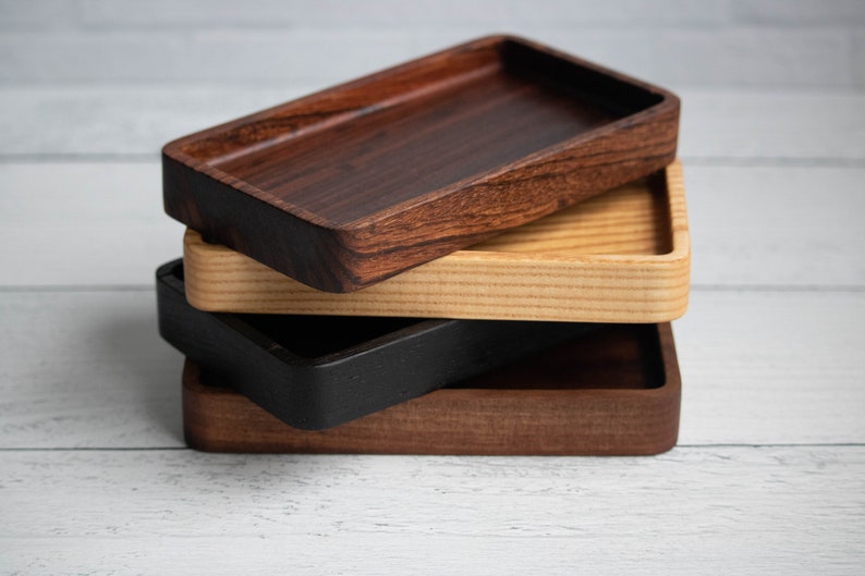 May include: A stack of four rectangular wooden trays in varying shades of brown and black. The top tray is a dark, rich brown, followed by a lighter wood, a black tray, and a medium brown tray at the bottom. The trays are stacked on top of each other.