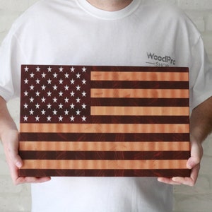 May include: A wooden cutting board shaped like the American flag. The board is made of maple and walnut wood and features a red, white, and blue design. The board is held by a person's hands.