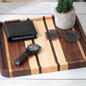 May include: A wooden tray with a walnut and maple striped design. The tray has a raised edge and is personalized with the initials "STE" and the date "11.04.2004". The tray is decorated with a black wallet, a watch with a black band and red stitching, and a pair of sunglasses.