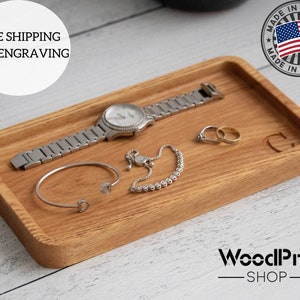 May include: A rectangular wooden tray with rounded edges, made of light brown wood. The tray is filled with jewellery, including a silver watch, a bracelet, and two rings. The tray has the letter "C" engraved on it.