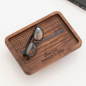 May include: A rectangular dark brown wooden valet tray featuring an engraved American flag design with a thin blue line. The tray is inscribed with the text Class 268 February 5, 2026. A pair of black-framed eyeglasses rests on the tray surface.