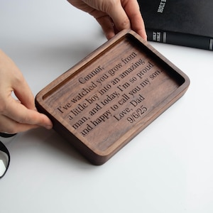 Puede incluir: Una bandeja rectangular de madera con acabado marrón oscuro. La bandeja tiene un interior empotrado y un borde elevado. El texto grabado dice: "Connor, I've watched you grow...Love, Dad 9/6/25."