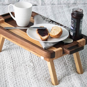 May include: A wooden breakfast tray with a white mug, a plate with a pastry and jam, and a jar of jam. The tray has a light wood base and a dark wood top with a handle on each side. The tray is sitting on a white fuzzy surface.