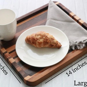Op de afbeelding: Een houten dienblad met een natuurlijke houten afwerking. Het dienblad is 35,56 cm lang en 25,4 cm breed. Het heeft een veelkleurig houten ontwerp met een lichtgrijze servet erop. Het dienblad is groot genoeg om een bord, een kopje en een croissant te houden.